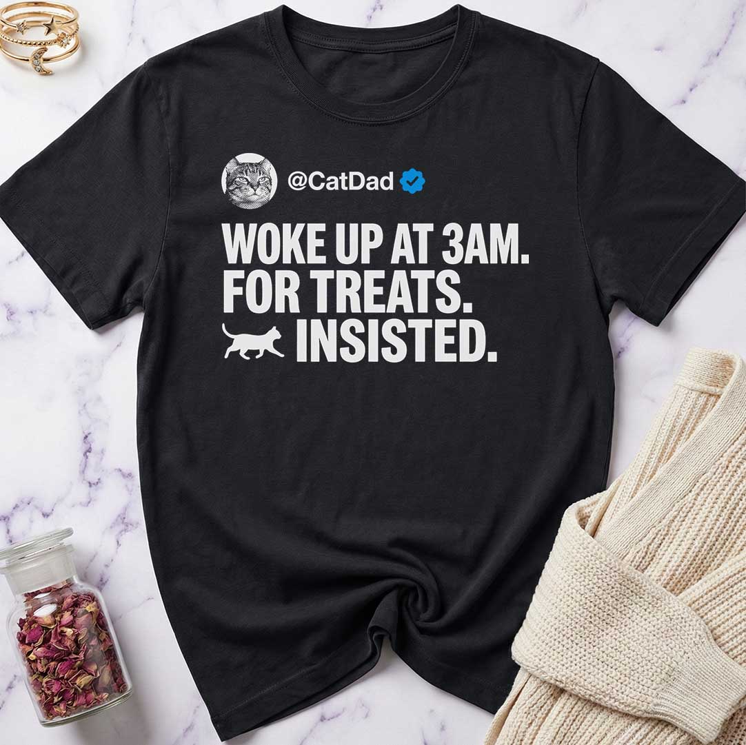 Black t-shirt with humorous text design on a marble surface with a sweater and jar of dried flowers.