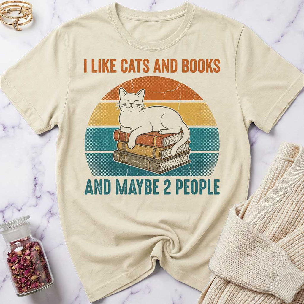 Beige t-shirt with a graphic of a cat on books and text 'I like cats and books and maybe 2 people' on a marble surface.