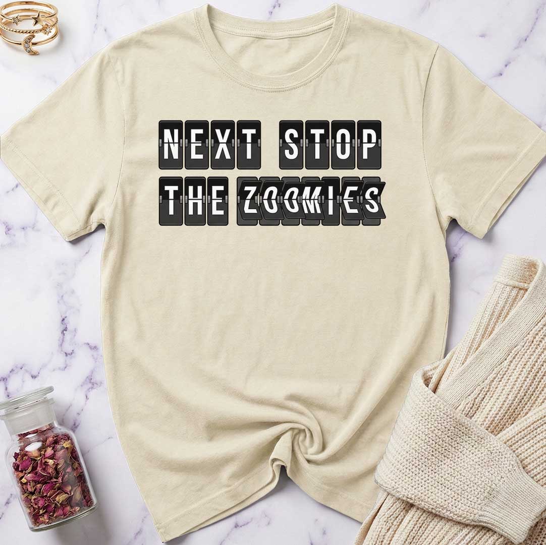Beige t-shirt with 'Next Stop The Zoomies' text on a marble surface with a sweater and jar of rose petals.