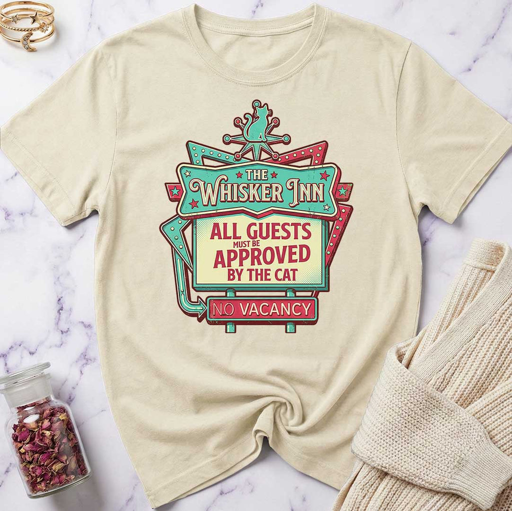 Beige t-shirt with 'The Whisker Inn' graphic design on a marble surface.