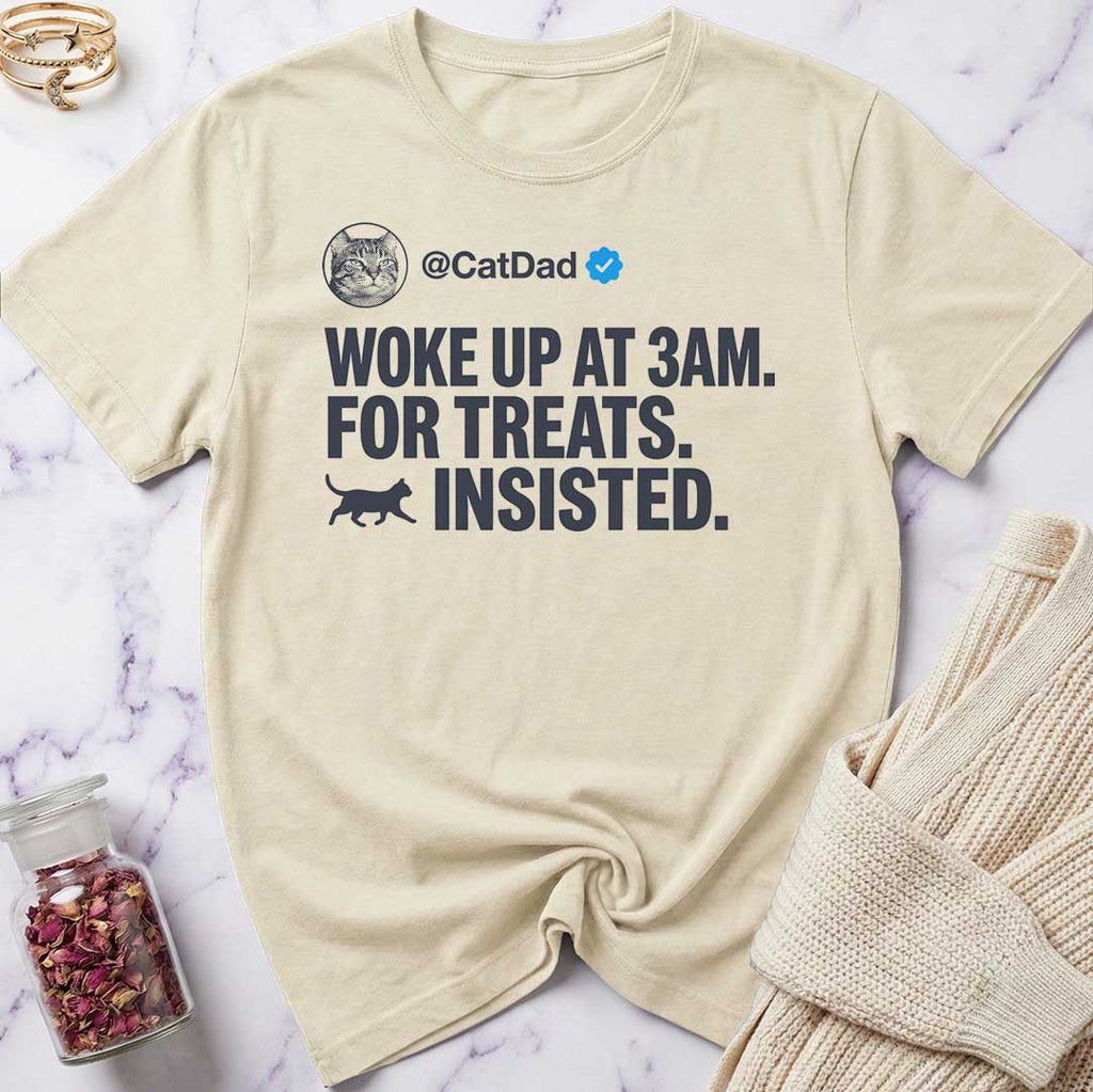 Beige t-shirt with humorous text design on a marble surface with a sweater and jar of treats.