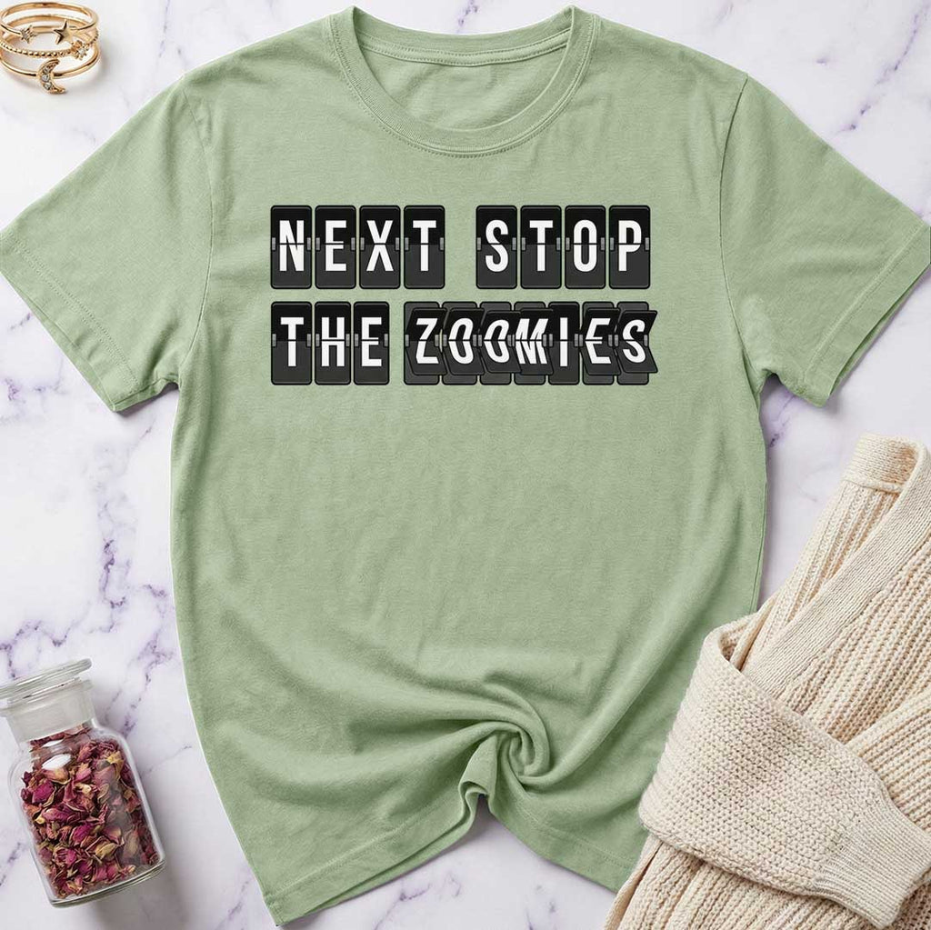 Green t-shirt with 'Next Stop The Zoomies' text on a marble surface with a beige sweater and jar of dried flowers.