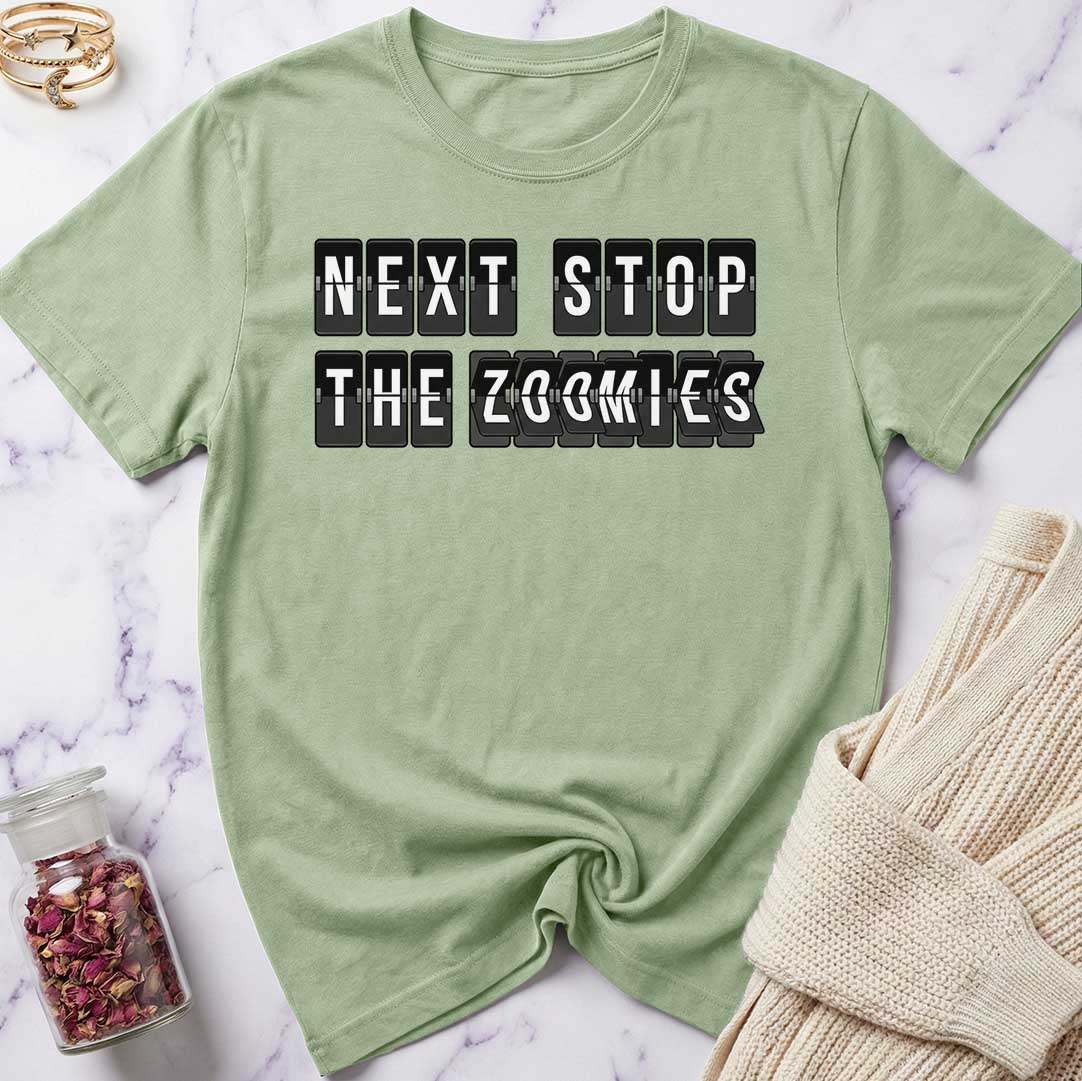Green t-shirt with 'Next Stop The Zoomies' text on a marble surface with a beige sweater and jar of dried flowers.